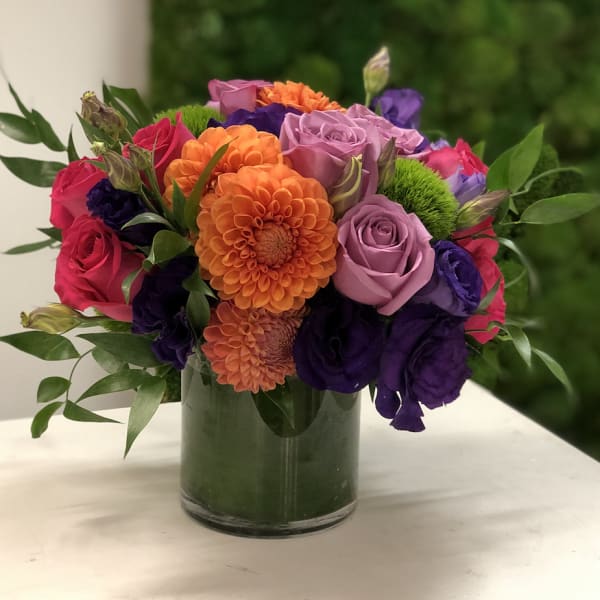 Compact arrangement of orange dahlias with purple and pink roses in a clear glass cylinder vase