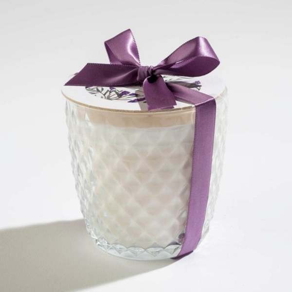 White candle in a textured glass jar with a floral lid and purple satin ribbon bow.