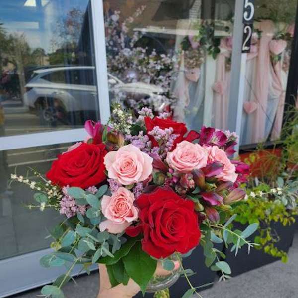 Small vase arrangement of red and pink roses with accent flowers, held in front of a shop window