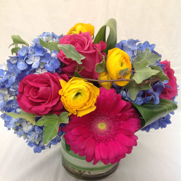 Bright arrangement of pink roses and gerbera daisy with yellow blooms and blue hydrangeas in a glass vase