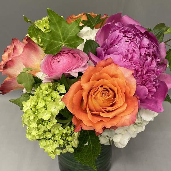 Compact arrangement of orange roses, pink blooms, and green hydrangea in a clear glass vase