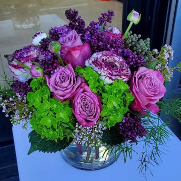 Compact arrangement of pink roses, purple blooms, and green hydrangeas in a clear glass vase