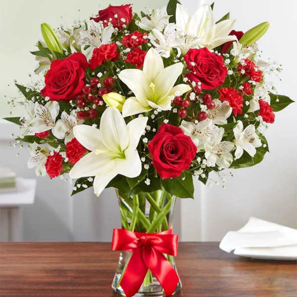 Arrangement of red roses, red carnations, and white lilies in a clear glass vase with a red ribbon bow
