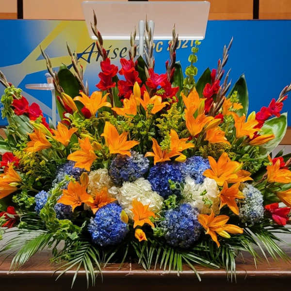 Large altar floral arrangement with orange lilies, blue and white hydrangeas, and red accent flowers on a stage