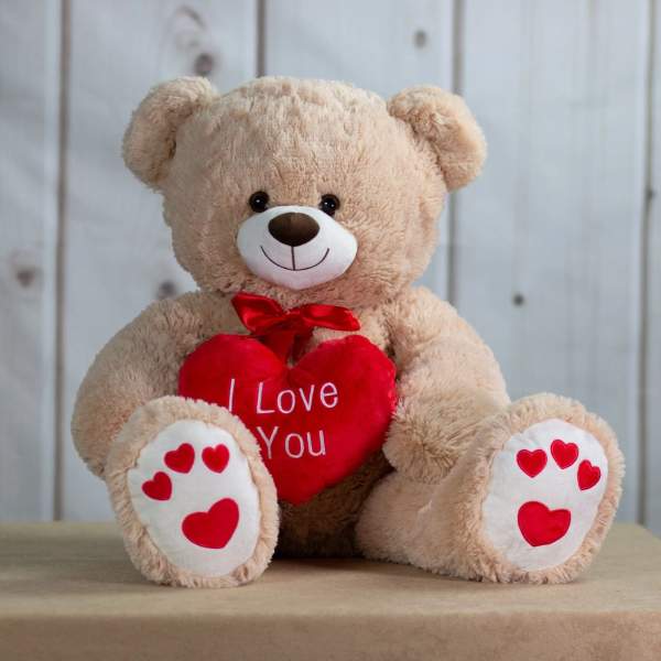Beige teddy bear holding a red heart that reads I Love You with red hearts on its paws.