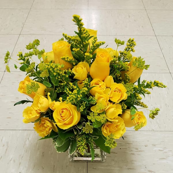 Yellow roses arranged in a clear square vase with green filler flowers