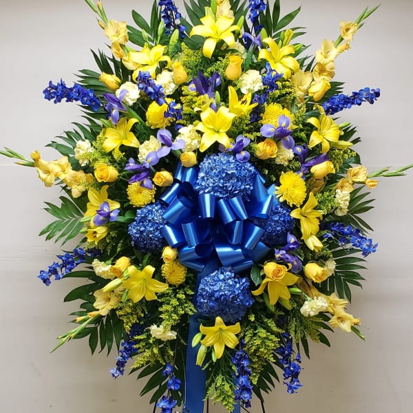 Large standing floral spray with yellow lilies, blue flowers, and a blue ribbon