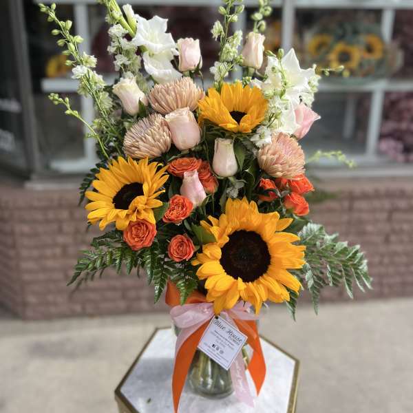 Bouquet of sunflowers, roses, and pale blooms in a glass vase with ribbons
