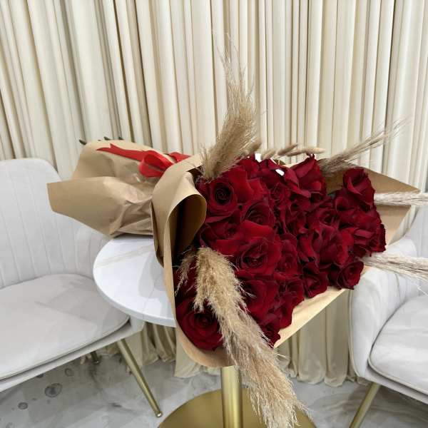 Large bouquet of red roses with pampas accents wrapped in brown paper on a small white table.