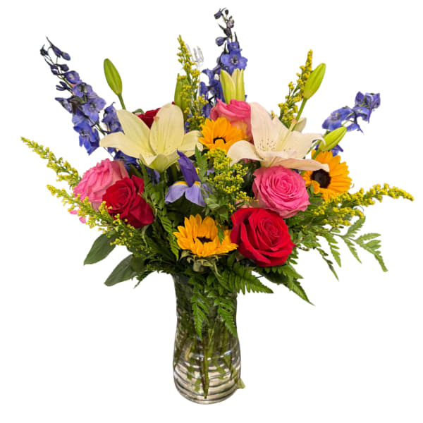 Mixed bouquet of roses, lilies, sunflowers, and blue delphinium in a glass vase
