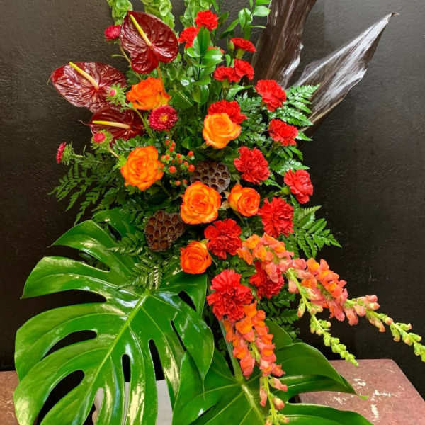 Tropical floral arrangement with orange roses, red carnations, and large monstera leaves