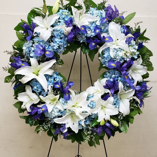 Standing open wreath of white lilies, blue hydrangeas, and purple iris on a metal easel.