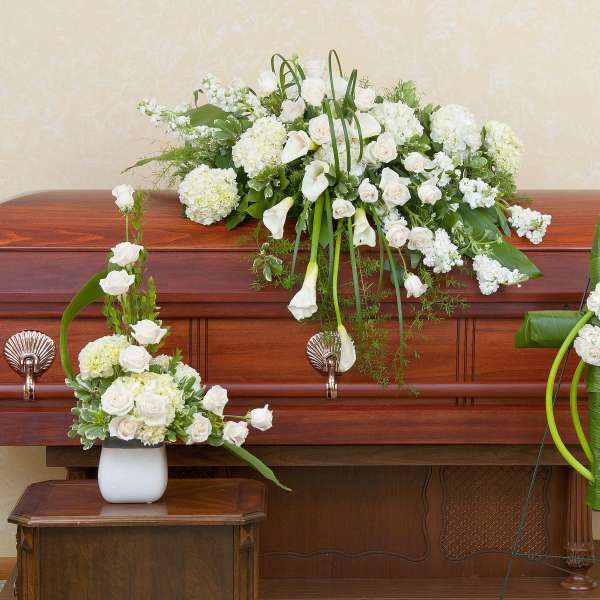 White funeral spray, vase bouquet, and cross arrangement displayed on a wooden casket