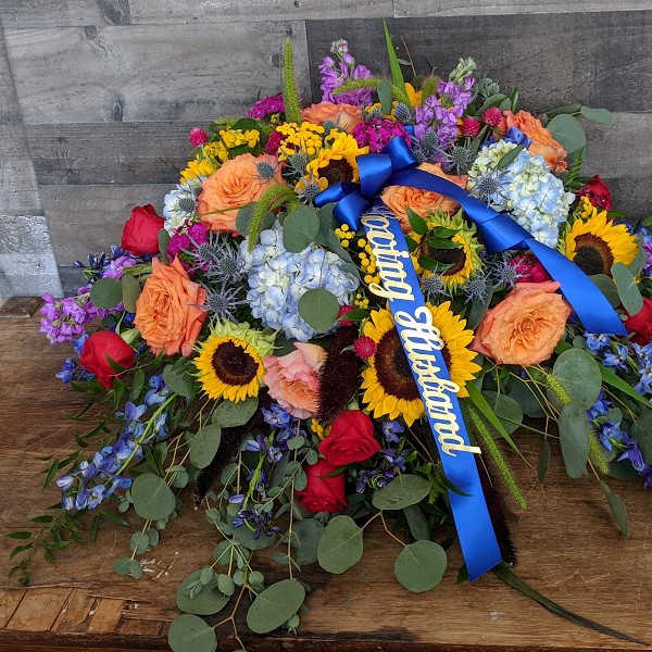 Colorful casket spray with sunflowers, roses, hydrangeas and a blue ribbon reading Loving Husband