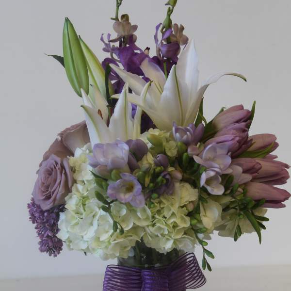 Mixed arrangement of white lilies, hydrangeas, and lavender blooms in a clear vase with a purple bow