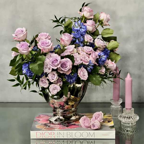 Lavender and pink rose arrangement with blue flowers in a silver vase beside pink candles