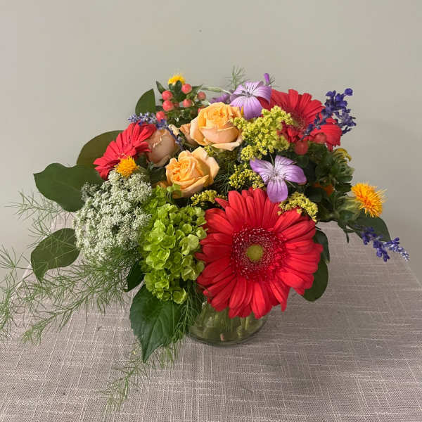 Mixed bouquet with red gerbera daisies, peach roses, and purple flowers in a glass vase