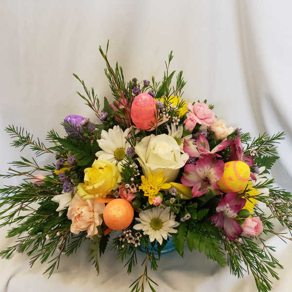 Low mixed spring arrangement with roses, daisies, alstroemeria and glitter Easter eggs in a blue container.
