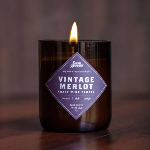 Lit wine-themed candle in a dark glass jar with a purple label on a wood surface
