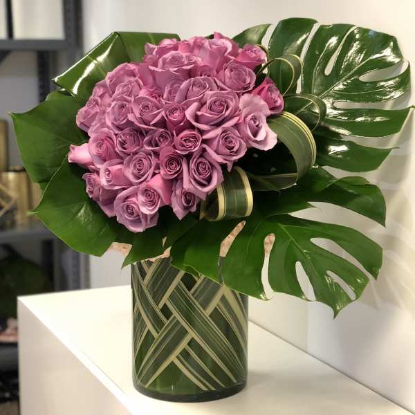 Tall modern arrangement of clustered lavender roses with large glossy leaves in a glass vase