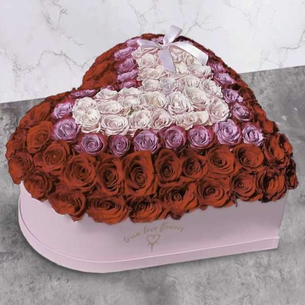 Heart-shaped boxed arrangement of red, pink, and lavender roses with a smaller pale heart and ribbon on top