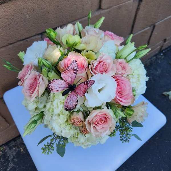 Round pastel arrangement of pink roses, white hydrangeas, and lisianthus with a pink butterfly decoration