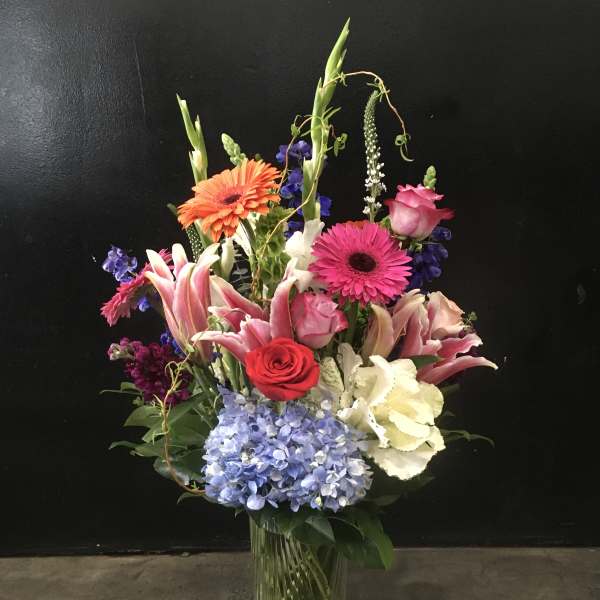 Tall mixed bouquet in a clear glass vase with pink, orange, blue, and white flowers.