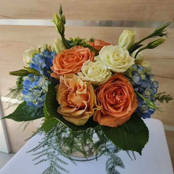 Compact arrangement of peach and cream roses with blue hydrangeas in a clear glass vase with feathery greenery.