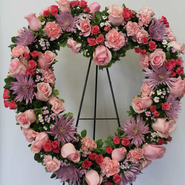 Heart-shaped wreath of pink roses, carnations, and lavender mums on a black metal easel.