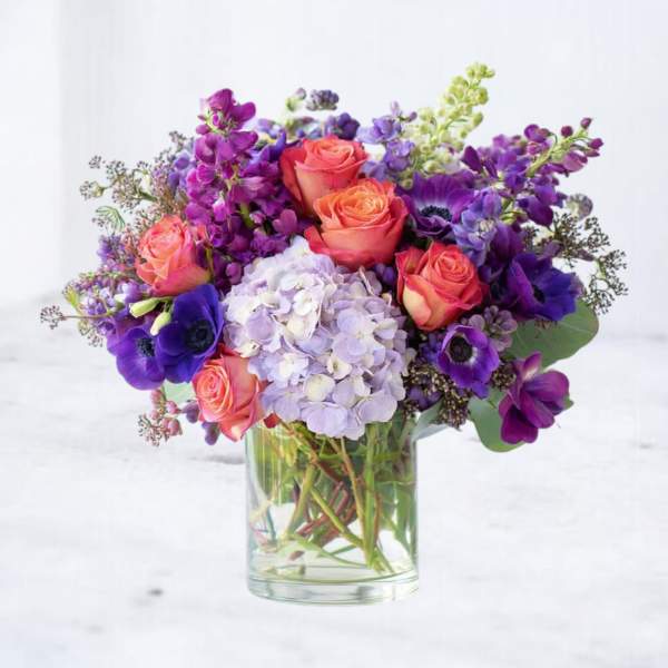 Mixed bouquet of peach roses, purple flowers, and lavender hydrangea in a clear glass cylinder vase