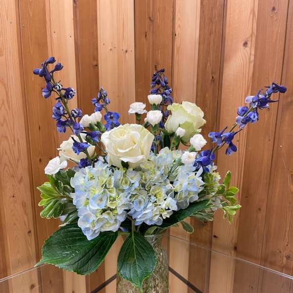 Blue hydrangeas with white roses and tall blue delphinium in a clear patterned glass vase