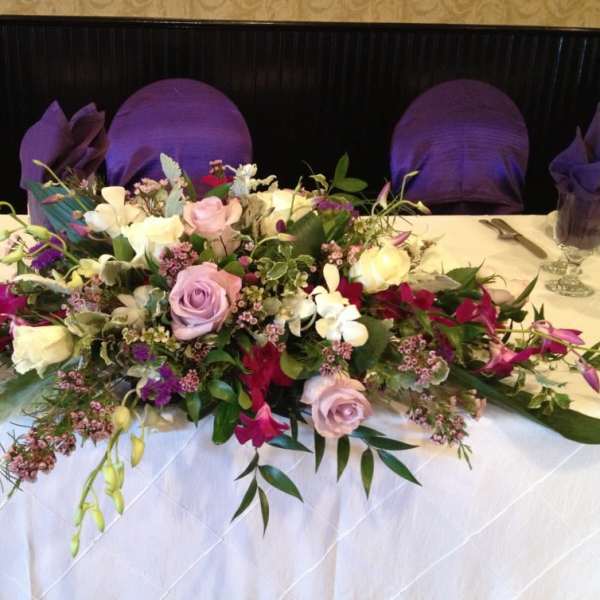 Low centerpiece of pink roses, white blooms, and purple flowers arranged across a banquet table