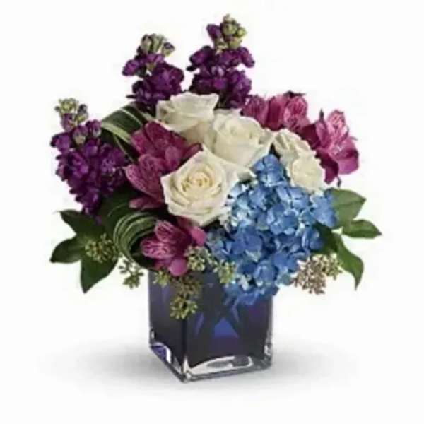 Jewel-toned arrangement with white roses, purple flowers, and blue hydrangeas in a dark square glass vase