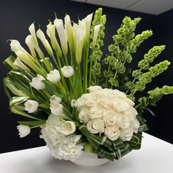 Tall white arrangement of calla lilies, roses, tulips and hydrangea in a low white bowl with green accents