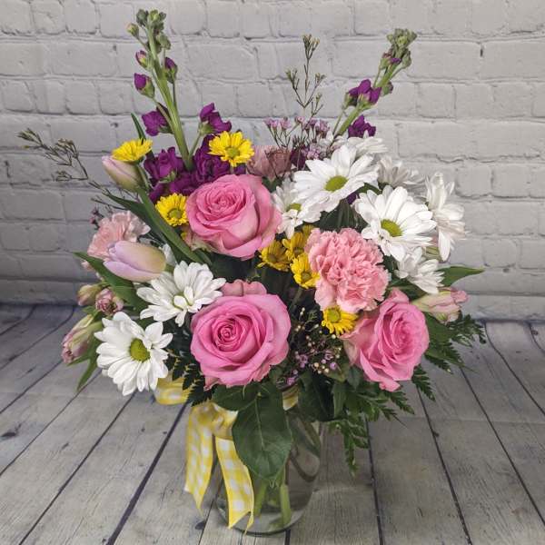 Pink roses, white daisies, and yellow flowers in a glass vase with a ribbon