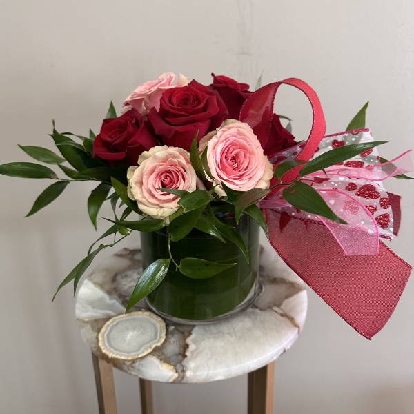 Short arrangement of red and pink roses with greenery in a glass vase, accented with a red and pink bow.