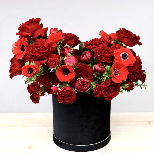 Lush arrangement of red roses, ranunculus, and anemones in a black round hatbox container
