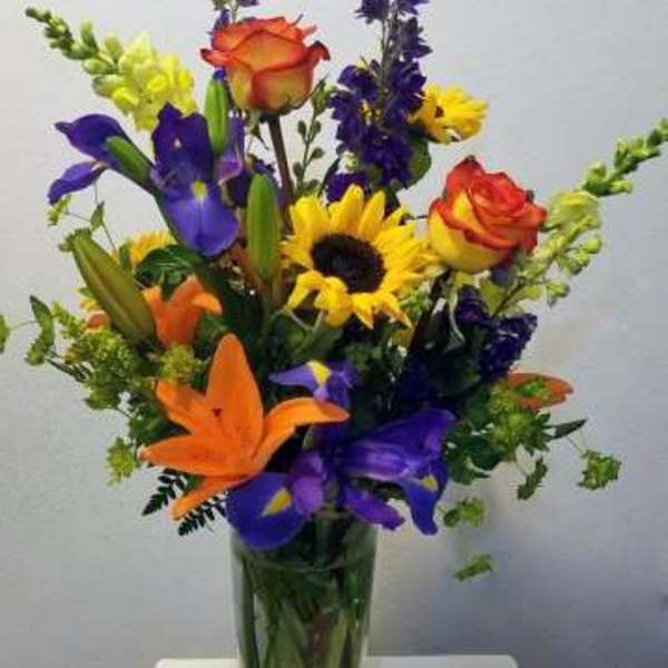 Tall mixed bouquet with orange lilies, yellow sunflowers, purple irises, and roses in a clear glass vase