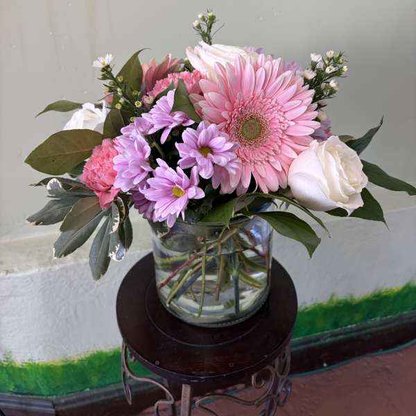 Pink gerbera daisies, roses, and lavender mums in a clear glass vase on a small stand