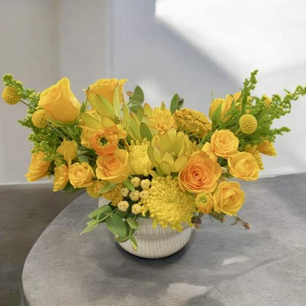 Low yellow flower arrangement with roses and other blooms in a white ceramic pot.