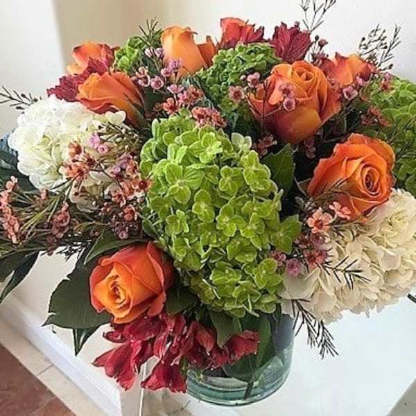 Low glass vase of orange roses, green and white hydrangeas, and red accent flowers.