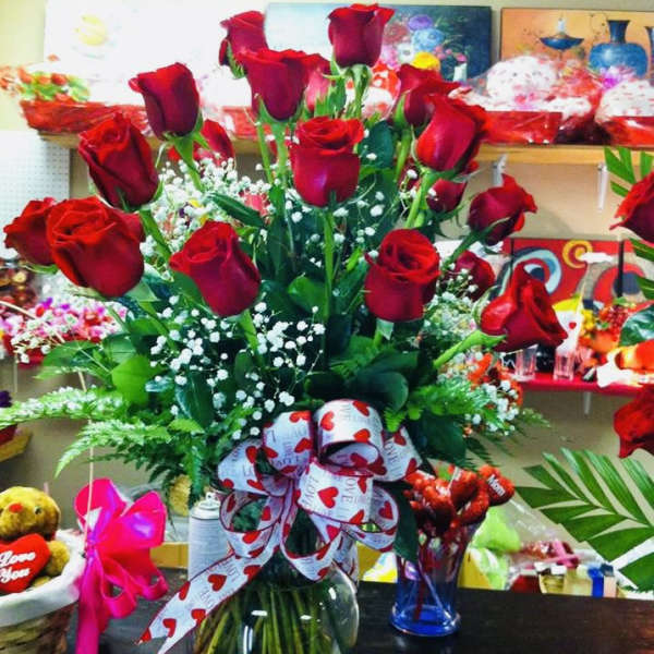 Tall arrangement of long-stem red roses with white filler flowers in a clear glass vase with a heart ribbon