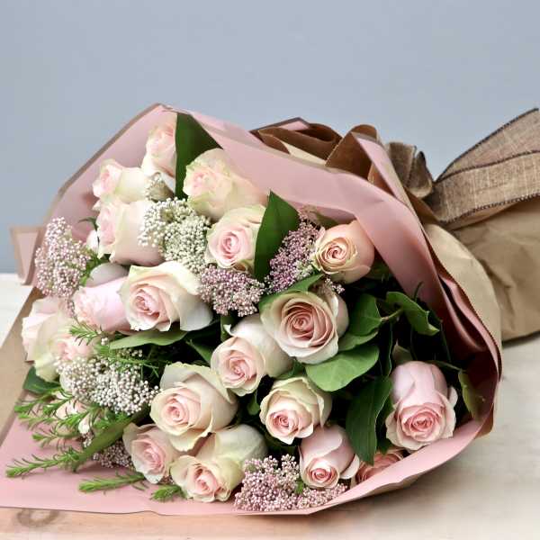 Hand-tied bouquet of pale pink roses with white filler flowers wrapped in pink and kraft paper