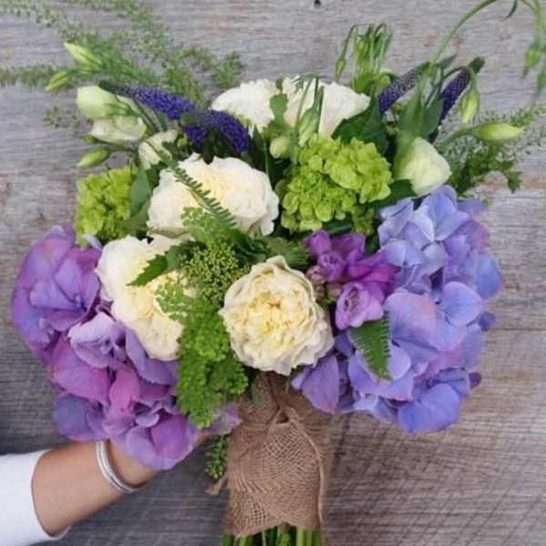 Hand-tied bouquet of cream blooms with purple hydrangeas and green accents wrapped in burlap