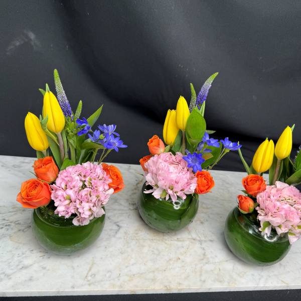 Three small floral arrangements in green glass vases with yellow, orange, pink, and blue flowers