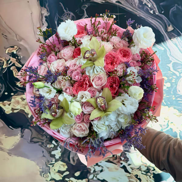 Round hand-tied bouquet of pink roses, white blooms, and green orchids wrapped in pink paper.