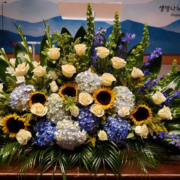 Large floral arrangement with white roses, blue hydrangeas, sunflowers, and purple spikes