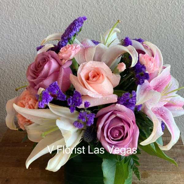 Compact arrangement of pink roses, lilies, and purple flowers in a green glass cube vase
