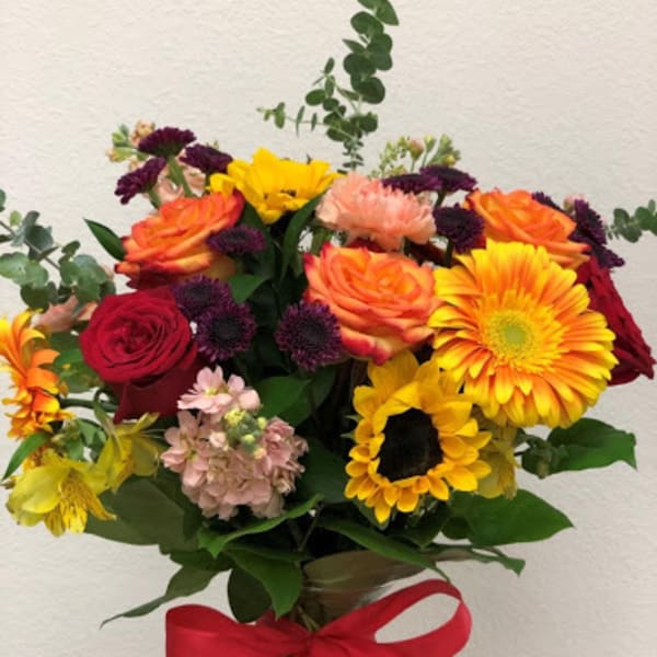 Mixed bouquet of roses, gerbera daisies, and sunflowers in a glass vase with a red ribbon