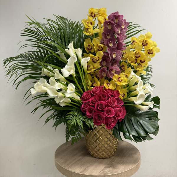 Tall tropical arrangement with pink roses, white calla lilies, orchids and large palm leaves in a gold vase